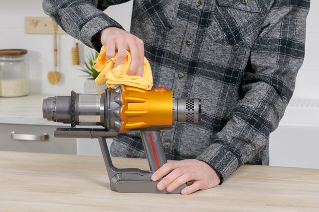 A person wearing a plaid shirt is cleaning an orange and gray handheld vacuum with a yellow cloth on a wooden table. The background includes kitchen items and a small potted plant.