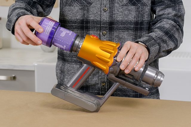 A person in a plaid shirt is assembling or disassembling a handheld vacuum cleaner on a wooden surface. The vacuum has a transparent dust container and orange details.