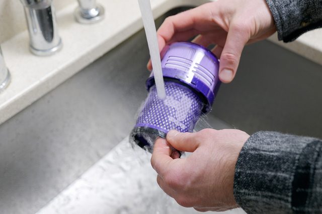 A person rinses a purple vacuum filter under a running tap in a sink. The person is wearing a dark plaid shirt. The water flows over the filter as it is being cleaned.
