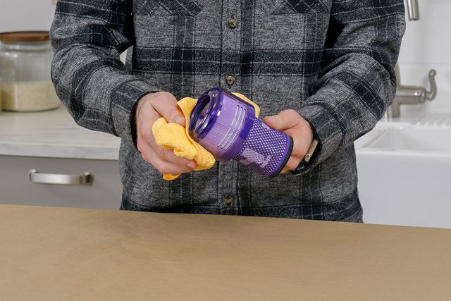 A person holding a yellow cloth and cleaning a purple vacuum cleaner filter over a countertop. The person is wearing a dark plaid shirt. A white sink and a jar can be seen in the background.