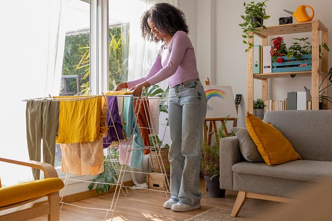 Smiling young woman drying clothes on rack by sofa in living room