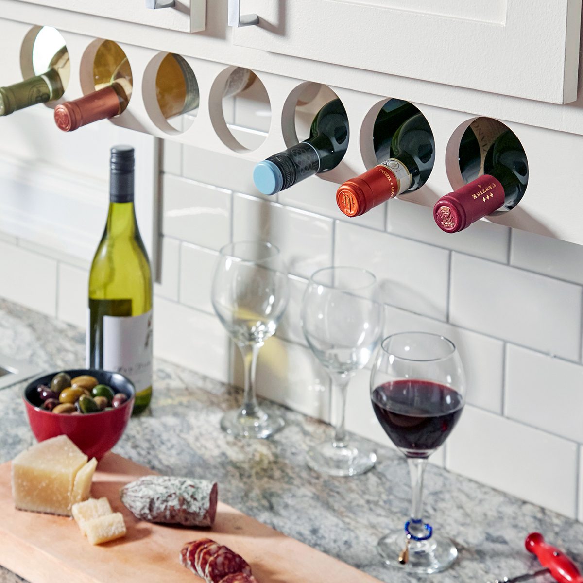 Six wine bottles are stored horizontally in circular cabinet slots above a kitchen counter with wine glasses, a bottle of white wine, a red wine glass, a bowl of olives, cheese, and sliced cured meats on a wooden board.