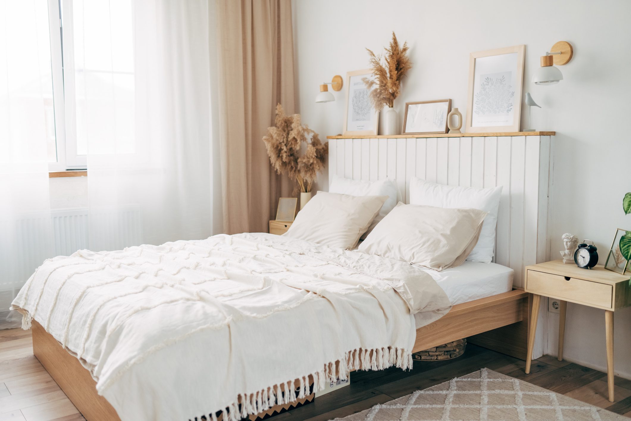 Simple bulb lamp on a rope hanging above bed with white bedclothes, books and gold fern leaf on an end table in white bedroom interior