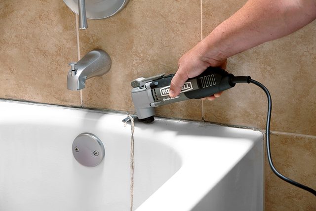 A person uses an electric oscillating multi-tool to remove old caulk from the edge of a white bathtub, next to tan tiled walls and a silver faucet.