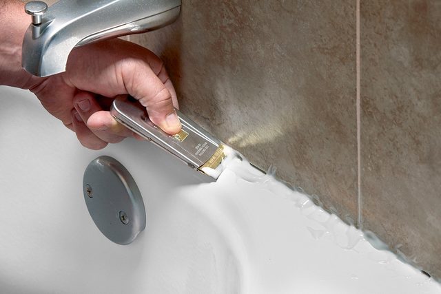 A hand uses a utility knife to remove old caulk from the edge where a bathtub meets a tiled wall near a faucet.