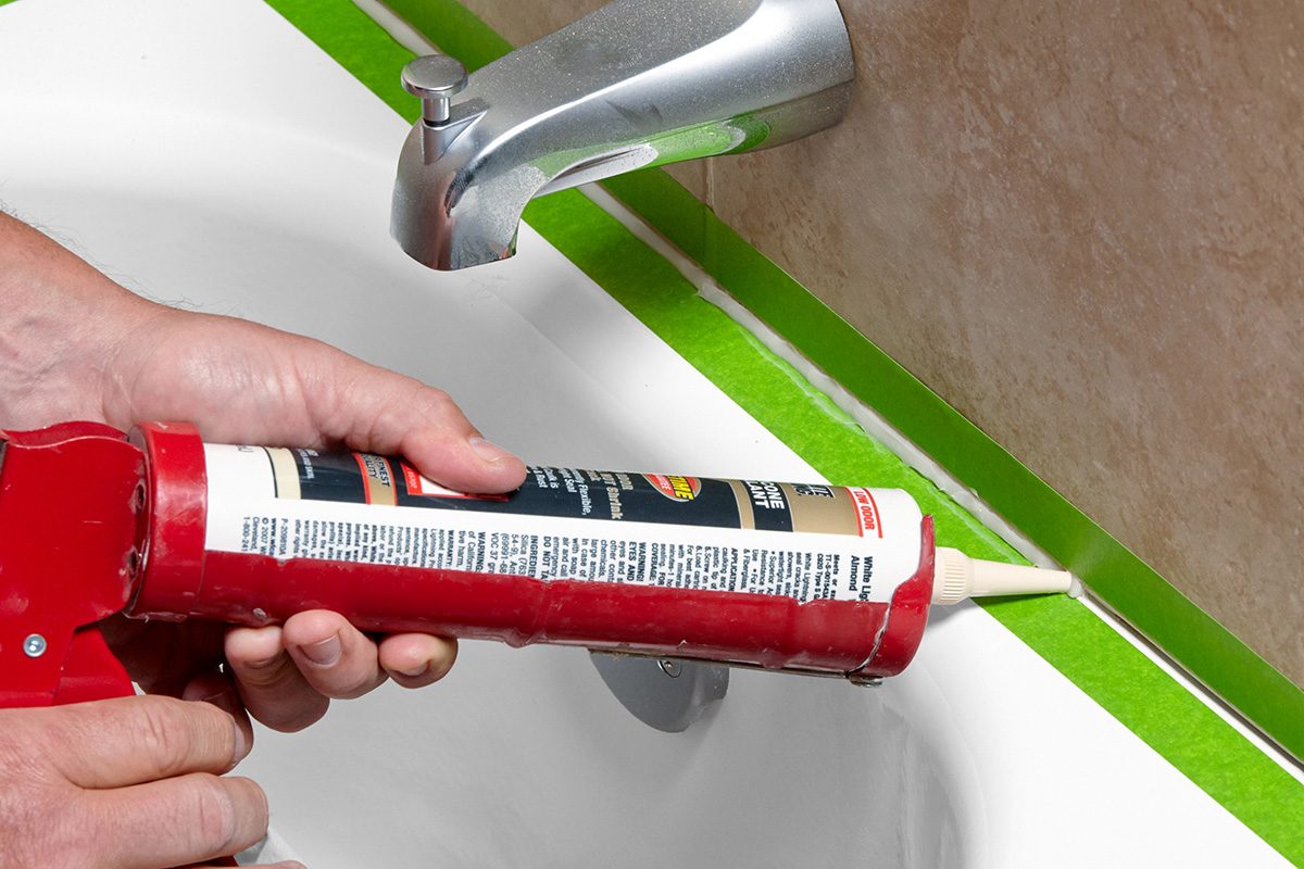 A person applies white caulk along the edge of a bathtub using a caulking gun. Green painter’s tape lines the seam between the tub and the tiled wall for a clean, precise finish.