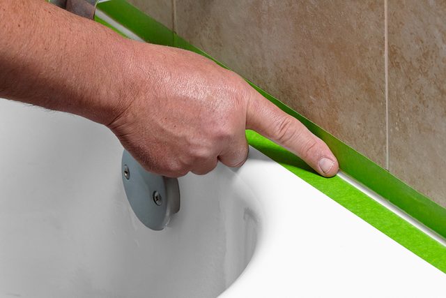 A person applies green painter’s tape along the edge where a white bathtub meets a tiled wall, preparing the surface, possibly for caulking or painting.