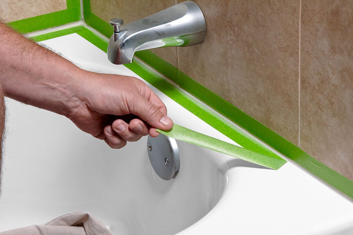 A person peels green painter’s tape from the edge of a bathtub, likely after painting or caulking, with a silver faucet and tiled wall visible in the background.