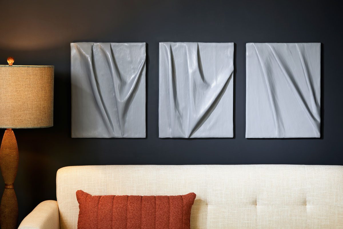 horizontal view of a cozy interior scene with a three section wall display and a sofa; the wall is painted a deep black, providing a striking backdrop for the three grey abstract panels that show folds and curves; to the left, a warm lamp with a beige shade; the cream colored sofa is accompanied by a rust colored throw pillow