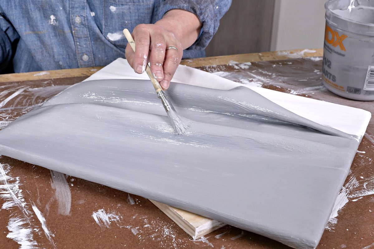 a person's hand applying gray paint to a canvas using a brush, the canvas is placed on a wooden surface, and there are streaks of paint and a small container of paint in the background;