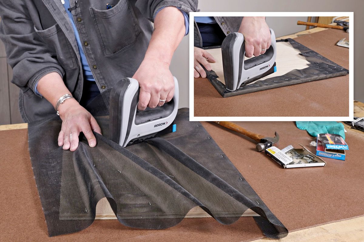 a person using a staple gun to attach a black mesh to a wooden surface; one hand holding the staple gun while the other holds the mesh in place; various tools visible in the background, including a hammer and a box of staples; a person using a staple gun to attach a fabric or material to a wooden board; the background includes a work table with various woodworking tools and materials; the surface of the table appears to be a brown colored material like fiberboard; there are some markings and light dust visible on the surface;