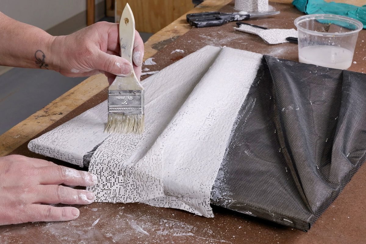 a person working on a canvas or fabric surface; the individual is applying a white substance with a brush; their hand is covered in paint or a similar material; the background features wooden textures, tools, and a container of liquid;