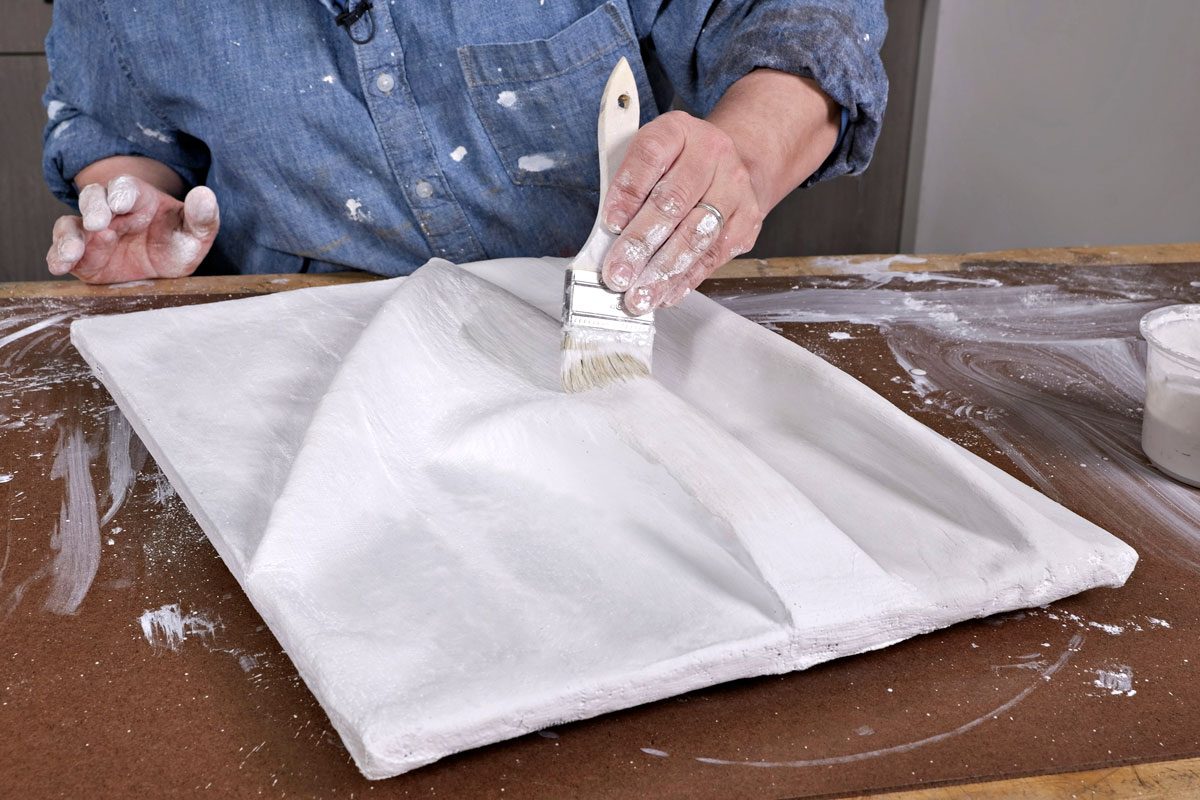 a person wearing a blue denim shirt, holding a paintbrush and applying a coat of white paint to a textured surface made of plaster; the surface has gentle folds, there are white paint splatters on the table surface, a small cup containing what looks like more white paint is also visible on the table,