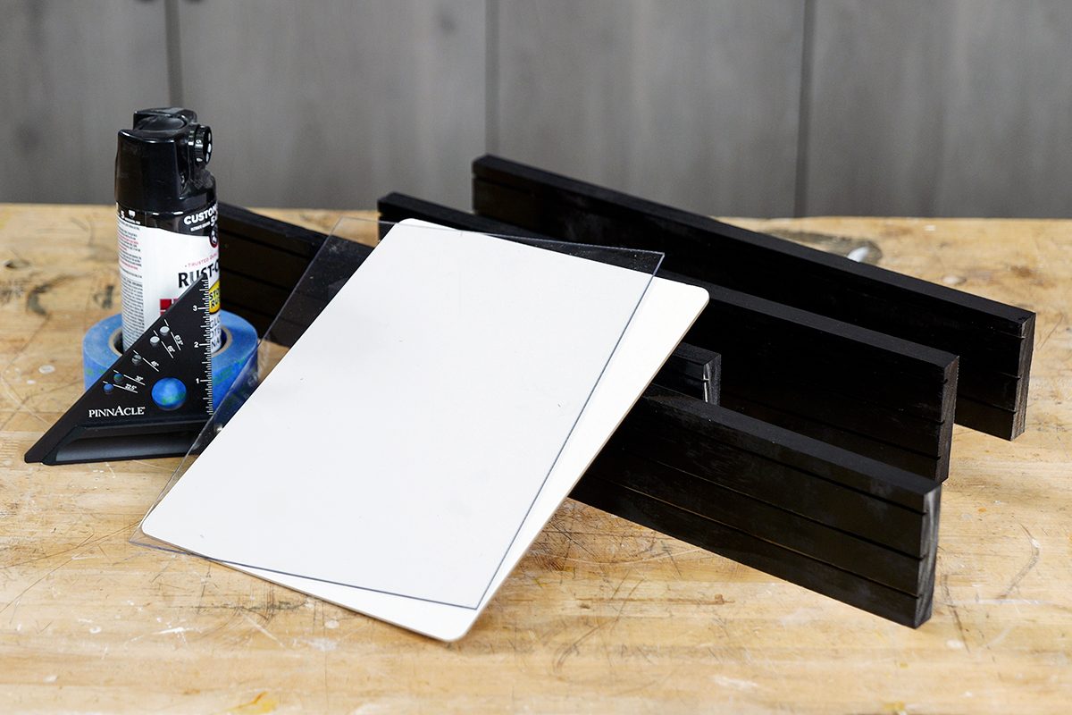 A glass cutter, black spray paint, blue tape, a rectangle sheet of glass, and a black wooden sled are arranged on a wooden workbench. A gray cabinet is visible in the background.