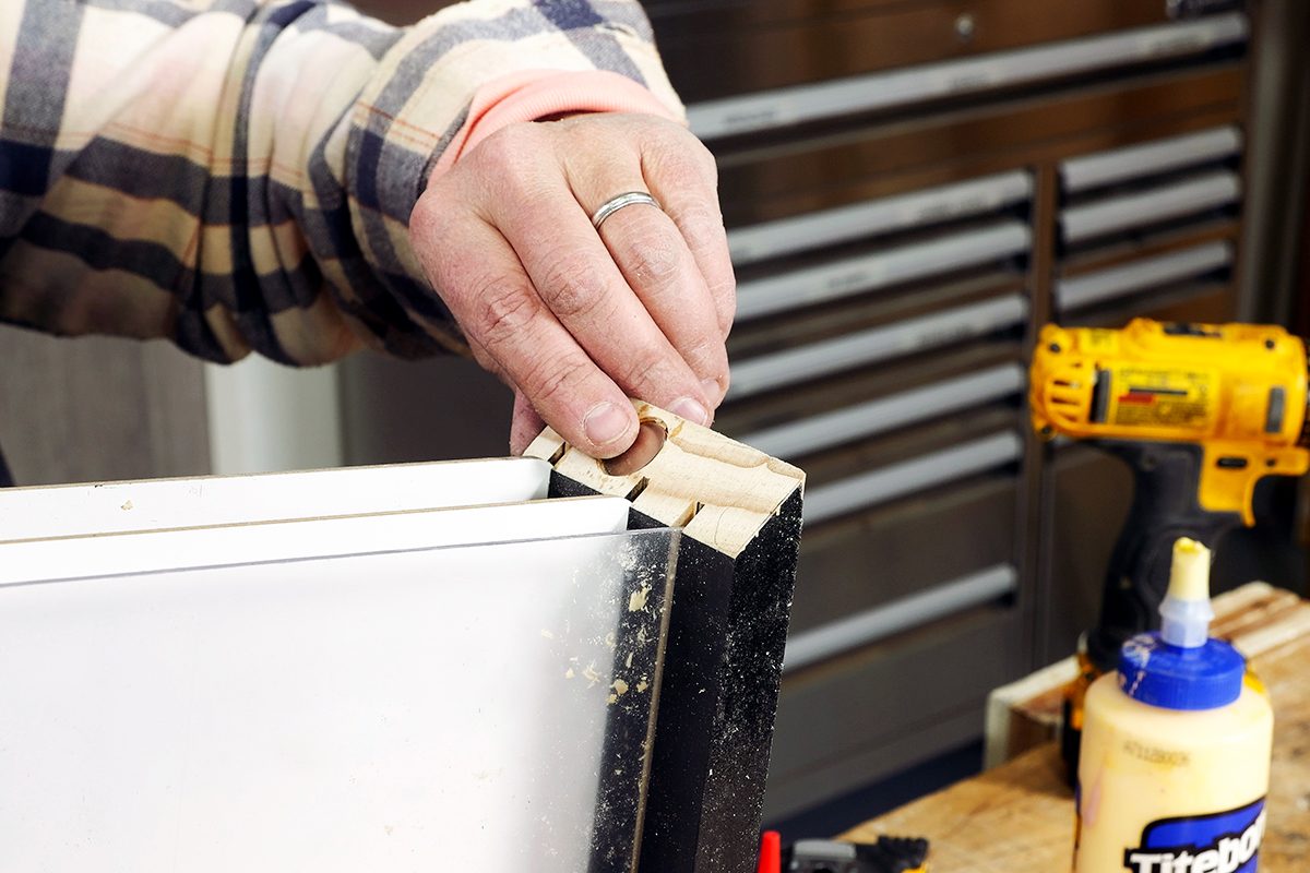 A person places wooden dowels into holes on the edge of a white cabinet panel. Nearby are a yellow drill, a bottle of wood glue, and a workbench, indicating a woodworking or furniture assembly project.
