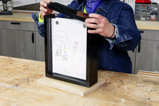 A person places a framed drawing with heart and stick figures onto a wooden base in a workshop, wearing a denim jacket and blue bandana.