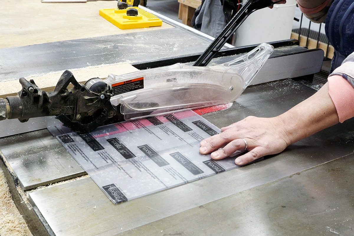 A person uses a table saw with a clear blade guard to cut a laminated sheet, keeping hands safely away from the blade in a woodworking shop.