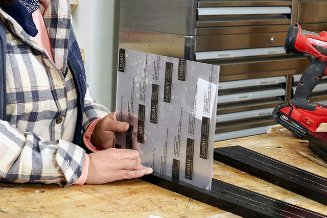 A person in a plaid jacket holds a rectangular metal sheet upright on a wooden workbench, with black metal strips and a red power tool nearby.