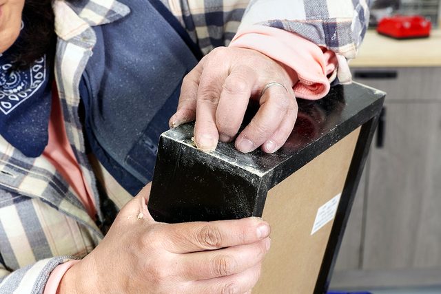 A person wearing a plaid shirt sands the corner of a black-framed object, possibly a piece of furniture or a picture frame, in a workshop setting.