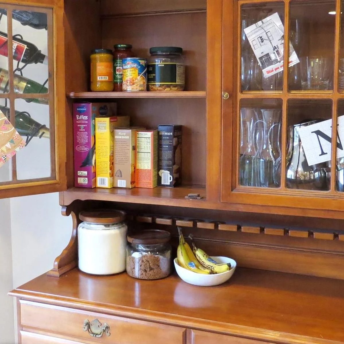 A wooden kitchen cabinet with glass doors displays glasses, cereal boxes, jars, bananas in a bowl, and containers of sugar and coffee on the countertop. There are notes and cards attached inside the cabinet.