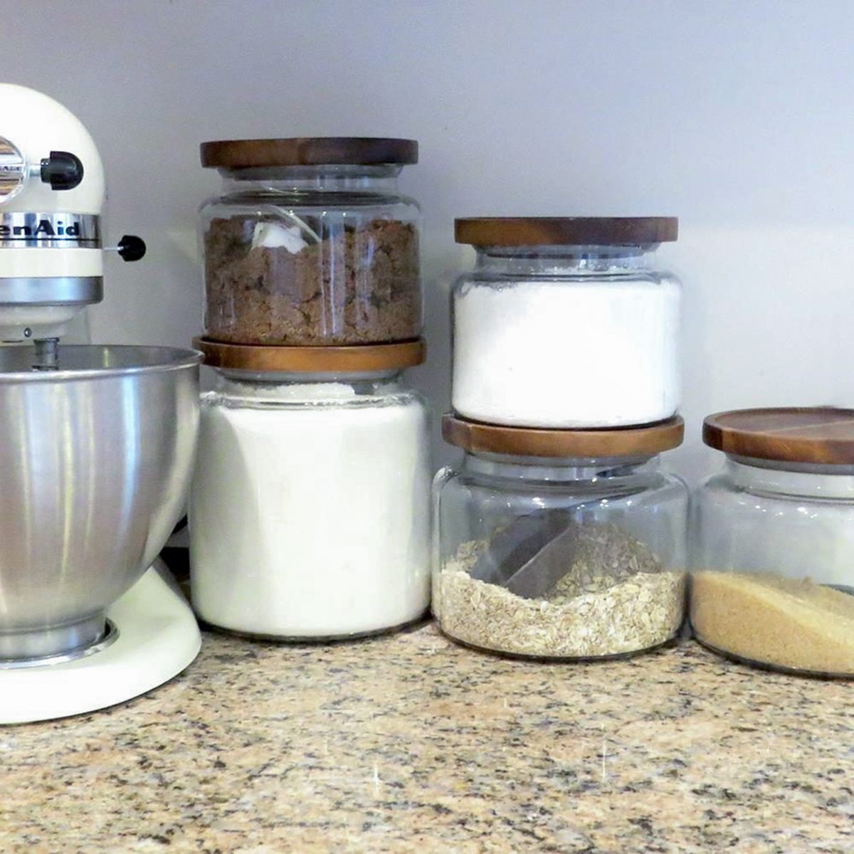 A kitchen countertop with a stand mixer and five glass jars with wooden lids, each filled with baking ingredients like flour, sugar, brown sugar, oats, and possibly cornmeal.