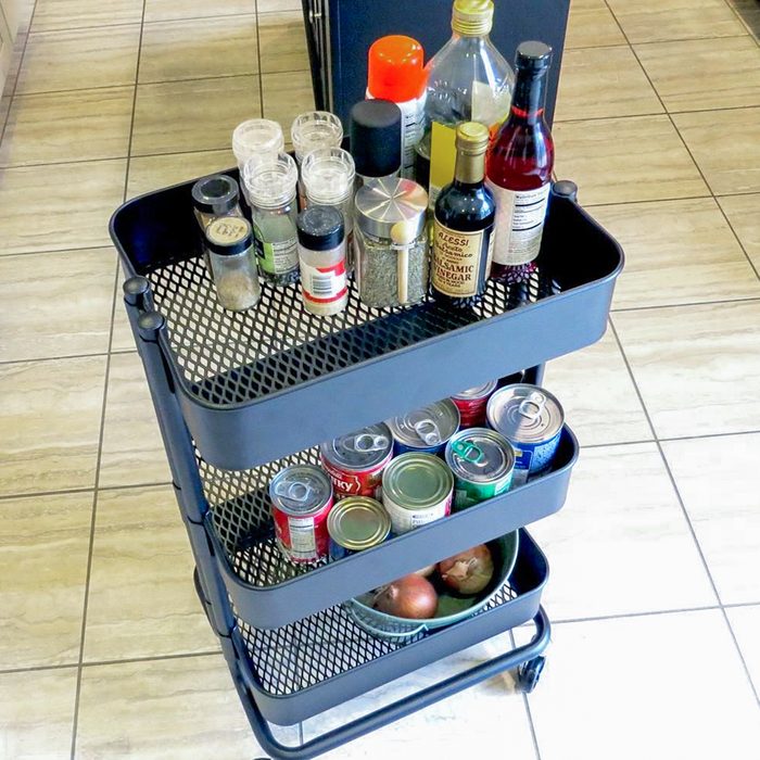 A black rolling utility cart with three shelves holding spices, condiments, canned drinks, and a bowl with onions, standing on a tiled floor.