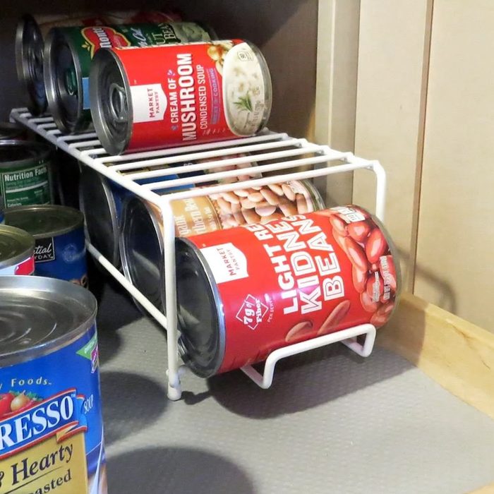 Cans of food, including light red kidney beans and cream of mushroom soup, are organized on a white metal rack inside a kitchen cabinet. Some other cans are visible to the side and underneath the rack.