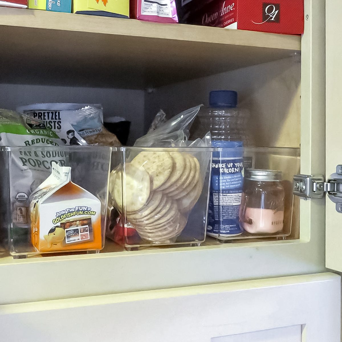A kitchen cabinet shelf holds clear plastic bins filled with snacks, including crackers, popcorn, a bottle, a jar, and a small carton, all neatly organized.