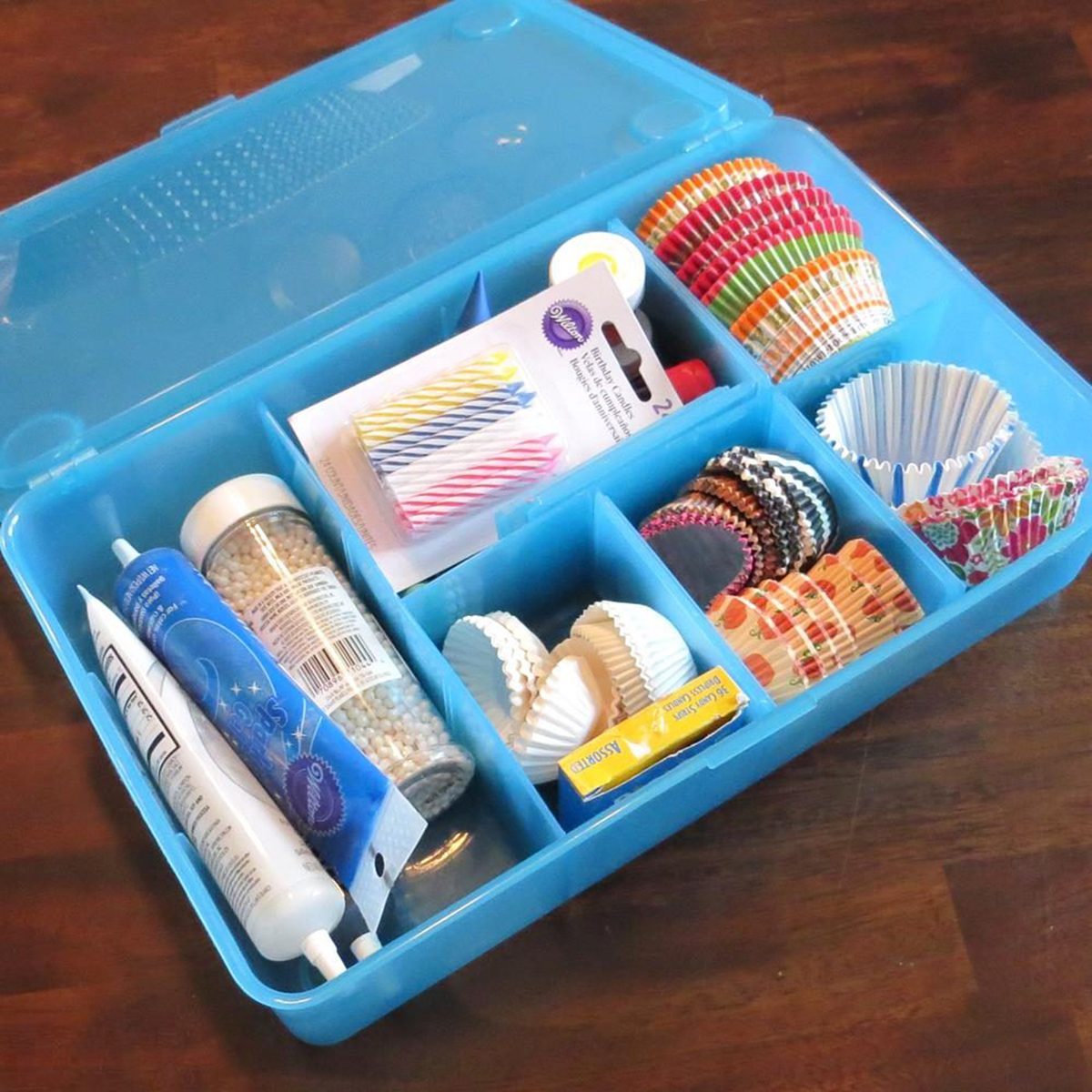 A blue plastic organizer box with compartments holding cupcake liners, birthday candles, sprinkles, icing tubes, and other baking supplies, placed on a wooden surface.