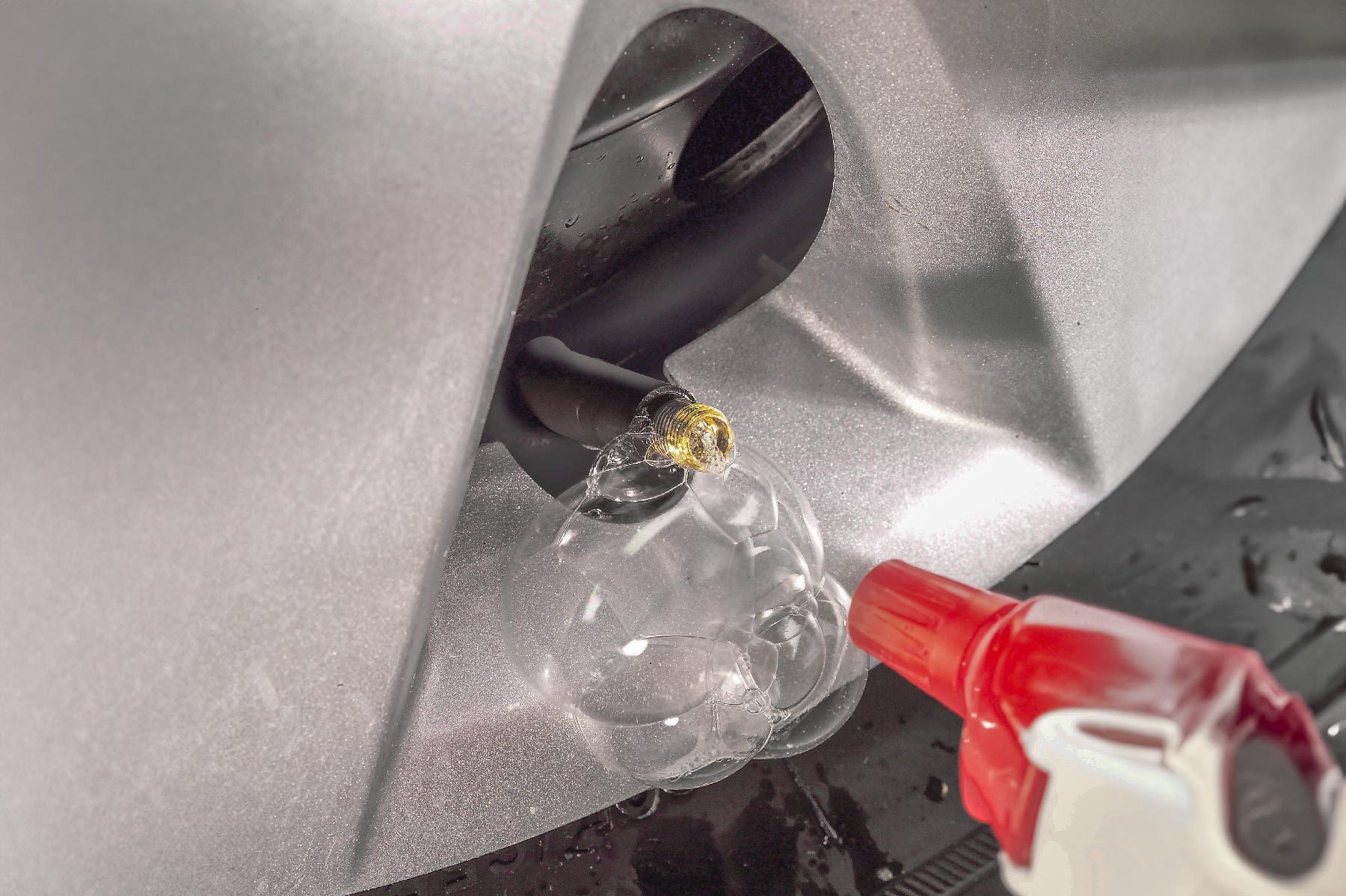 A clear bubble forms at a connection point where a hose extends from a silver surface, while a red spray nozzle directs liquid towards it.