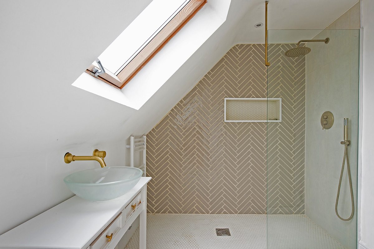 A general interior view of a white bathroom, glass bowl sink on a shabby chic dresser, towel radiator, brushed gold effect tap, skylight, hexagon tiled floor, herringbone patterned tiled wall, drench shower head and glass partition in a loft extension within a home