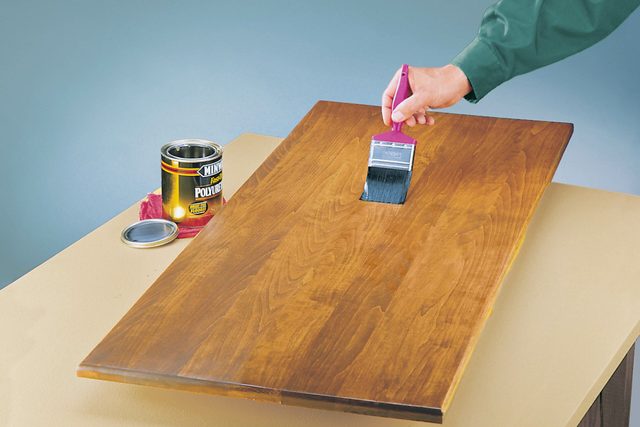 A hand applies a brush to a wooden surface, coating it with finish. Nearby are a can of polyurethane and a cloth, set on a yellow table.