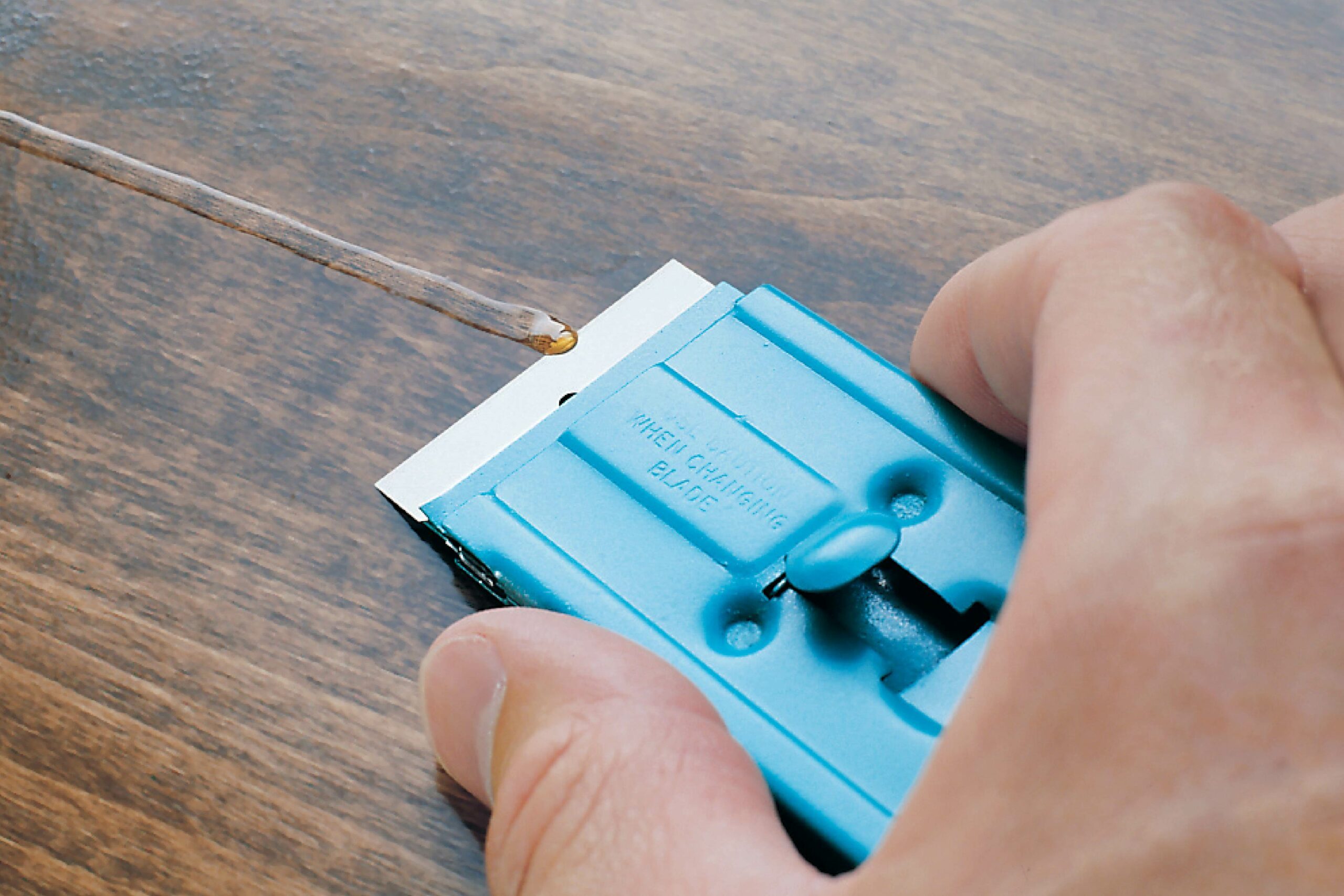 A hand holds a blue scraper, removing a thin line of adhesive from a wooden surface, revealing its smooth texture beneath.