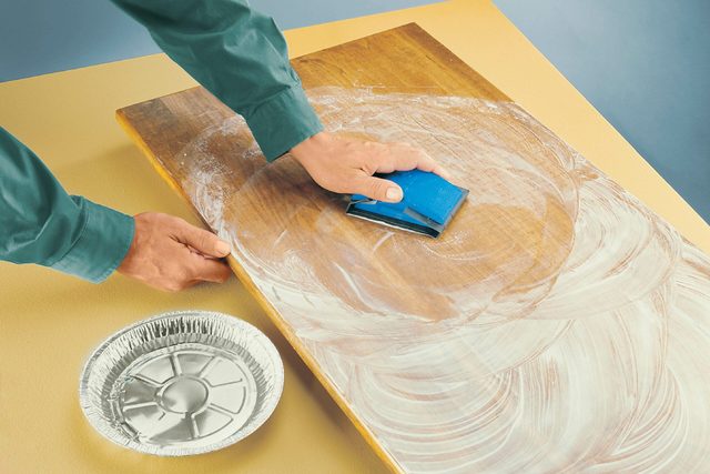A hand scrubs a wooden surface with a blue cleaning tool, while a round aluminum dish sits nearby, all set against a vibrant backdrop.