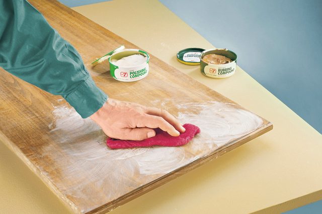 A hand applies red polishing cloth to a wooden surface, removing white polish, with two containers of polishing compound nearby on a vibrant tabletop.