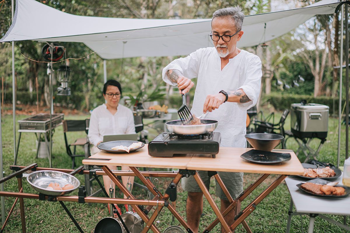 Asian chinese senior couple at camping tent, senior man cooking and senior woman using laptop