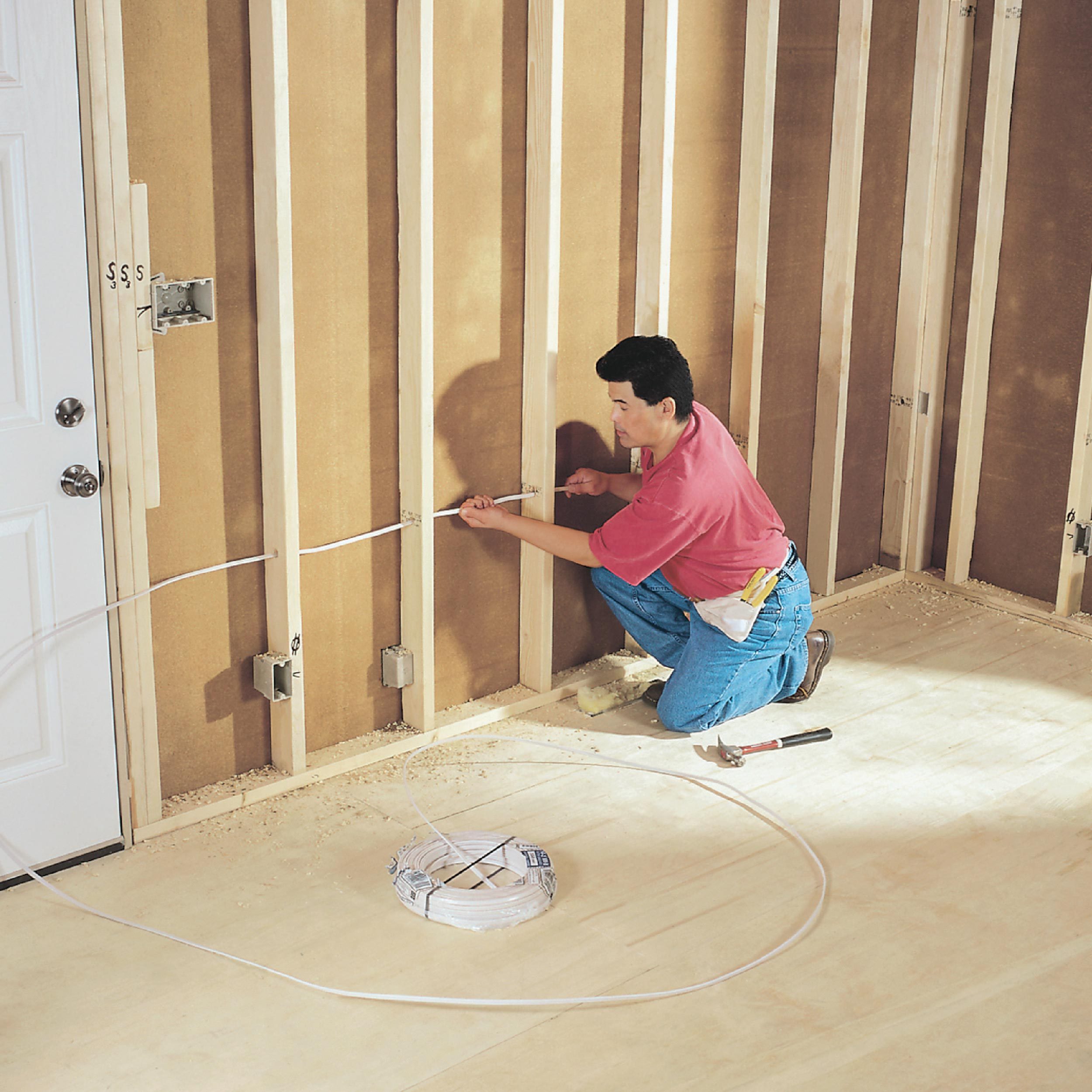 A man kneels, installing wiring on wooden wall studs in a partially constructed room, surrounded by framing and tools on the floor.
