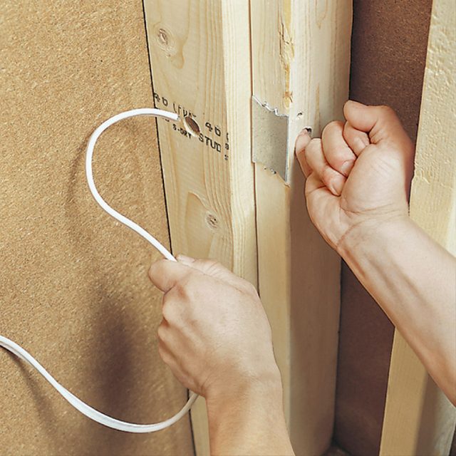 A hand pulls an electrical cord through a wall opening, while another hand secures the cord against wooden studs in a home improvement setting.