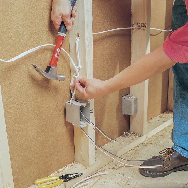 A hand holds a hammer, striking a wall. Another hand manipulates electrical wires near an electrical box in a wooden-framed interior space.
