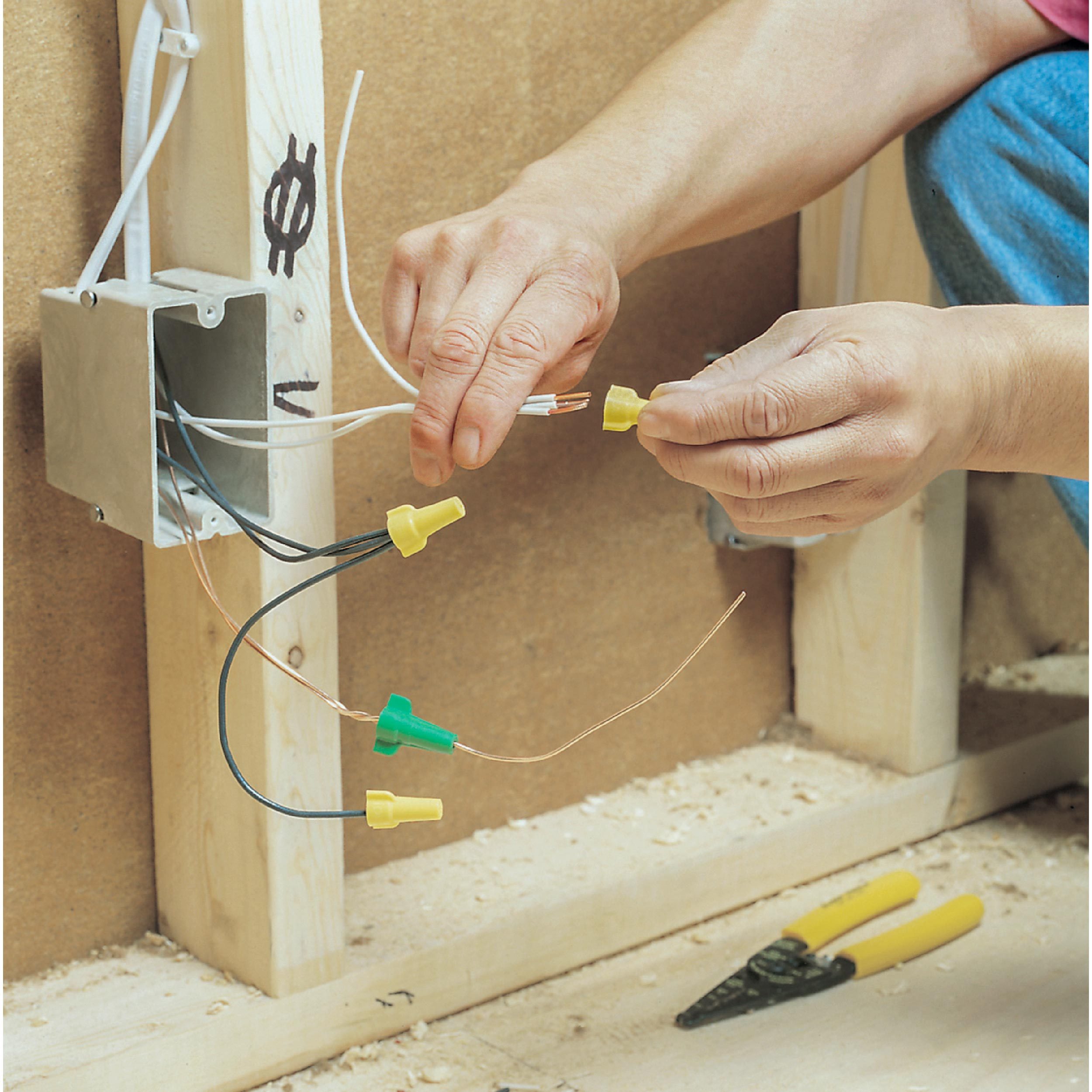 A person connects electrical wires using wire nuts, with an exposed electrical box mounted on a wooden framework in a construction setting.