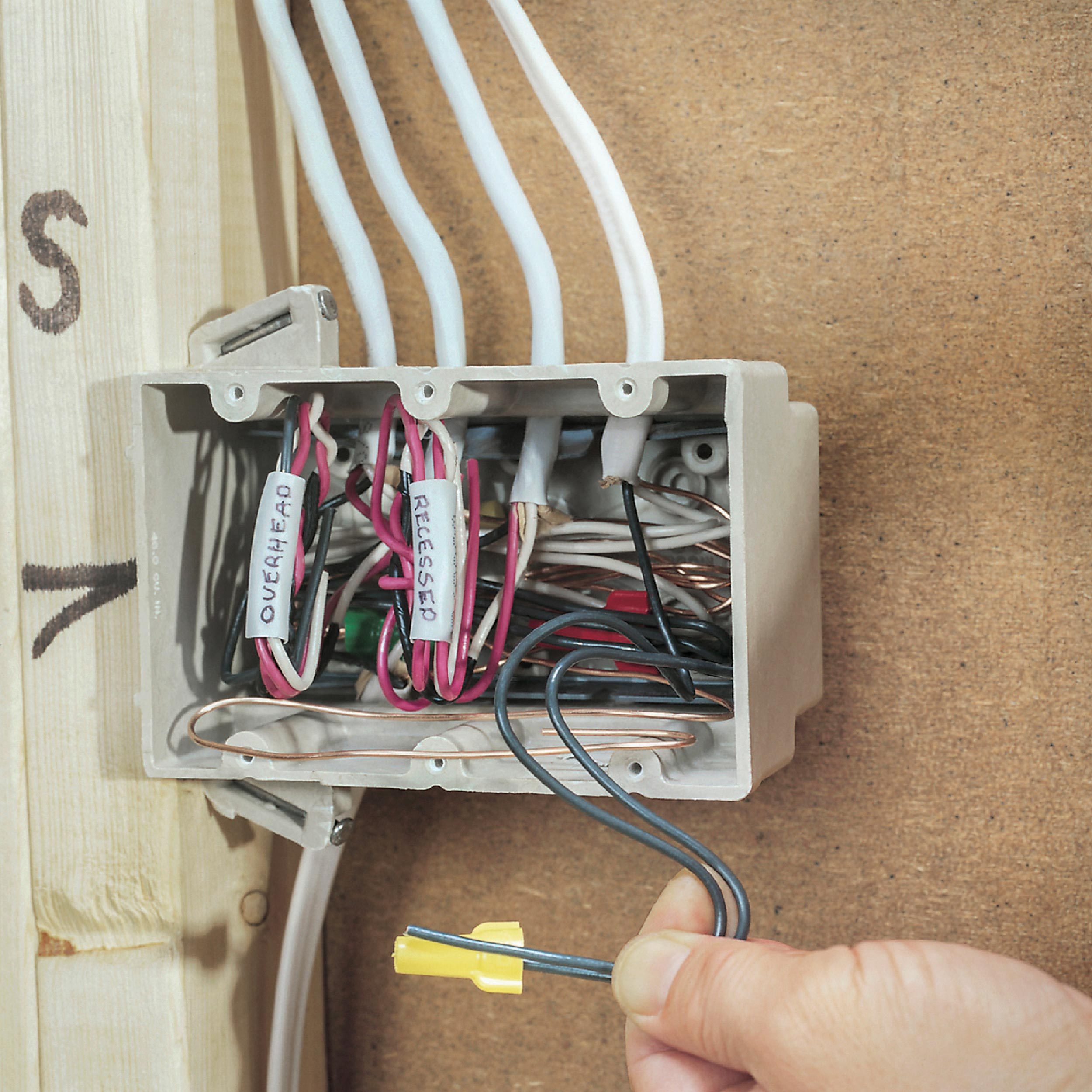 A hand holds a black wire near an electrical junction box filled with labeled wires, mounted against a wooden background.