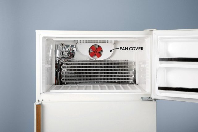 An open refrigerator interior features a red fan behind a cover and metal coils for cooling, set against a grey wall.