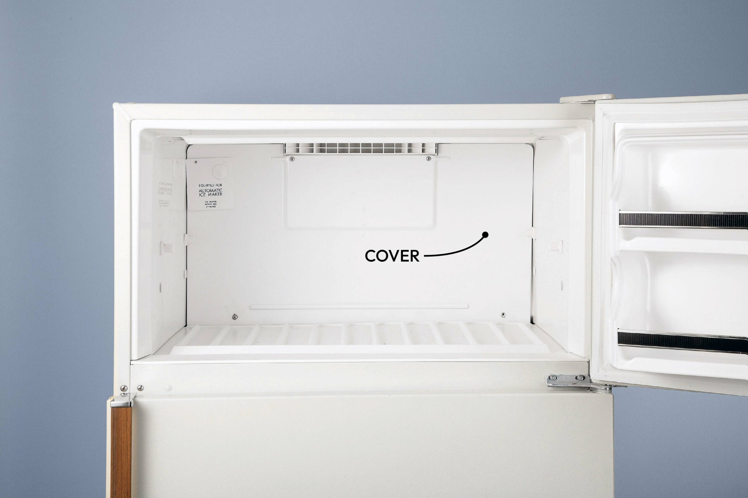 An open freezer reveals a white interior with an arrow pointing to a cover. The background is a solid grey wall.
