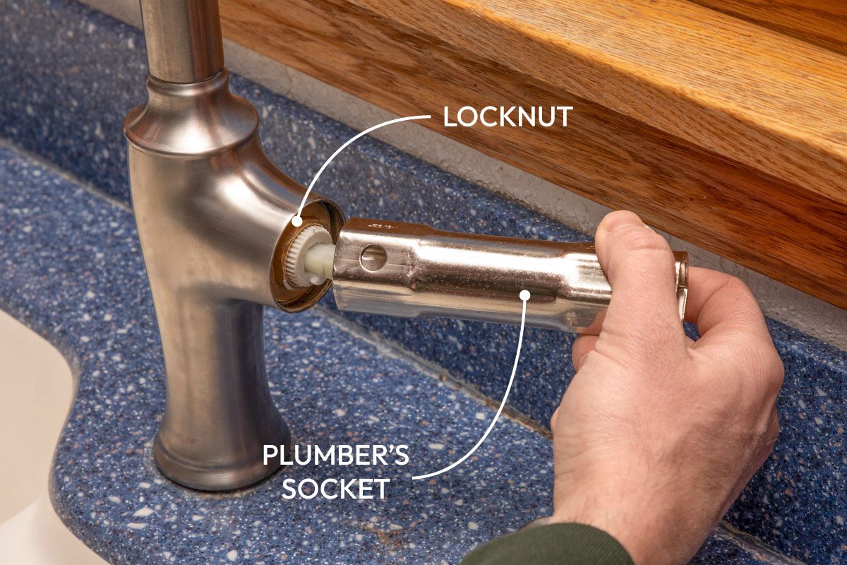 a hand holding a shiny metal cylindrical piece with hexagonal openings and inserting it into the side of a brushed metal kitchen faucet; a white plastic part is visible inside the faucet opening; the faucet is mounted on a blue speckled countertop, and a blurred wooden surface is in the background