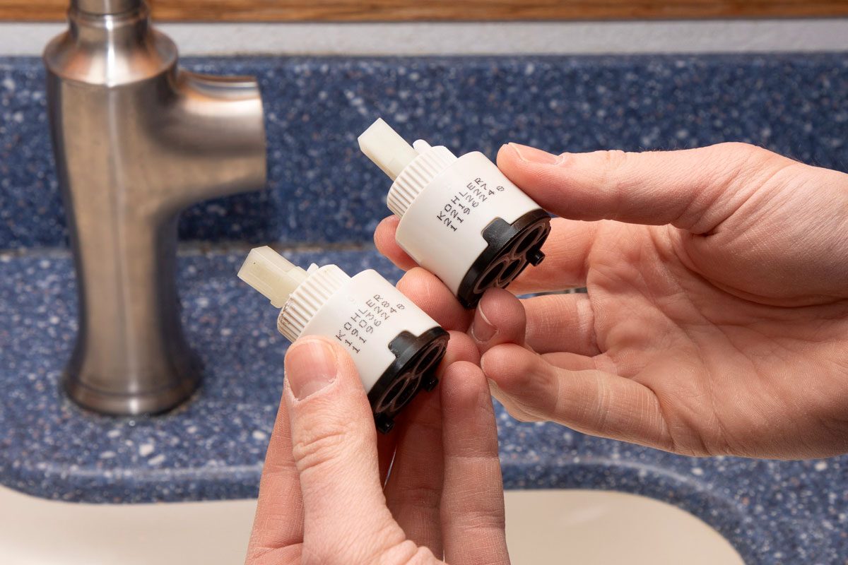 two hands holding two identical white cylindrical cartridges; in the blurred background, a brushed metal kitchen faucet is visible mounted on a blue speckled countertop with a white sink