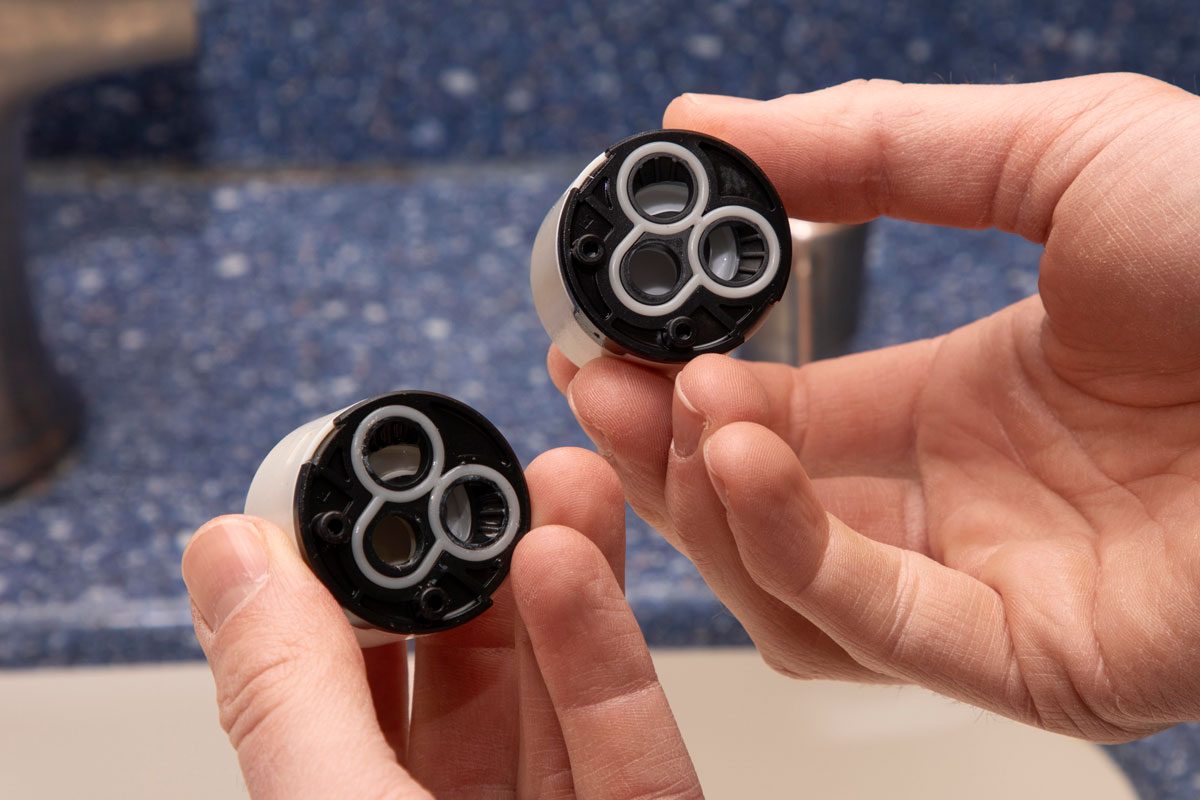 two hands holding two identical faucet cartridges; the cartridges are cylindrical with a white plastic body and a black bottom featuring three openings with white rings around them; the background is blurred but shows a blue speckled countertop and a brushed metal faucet