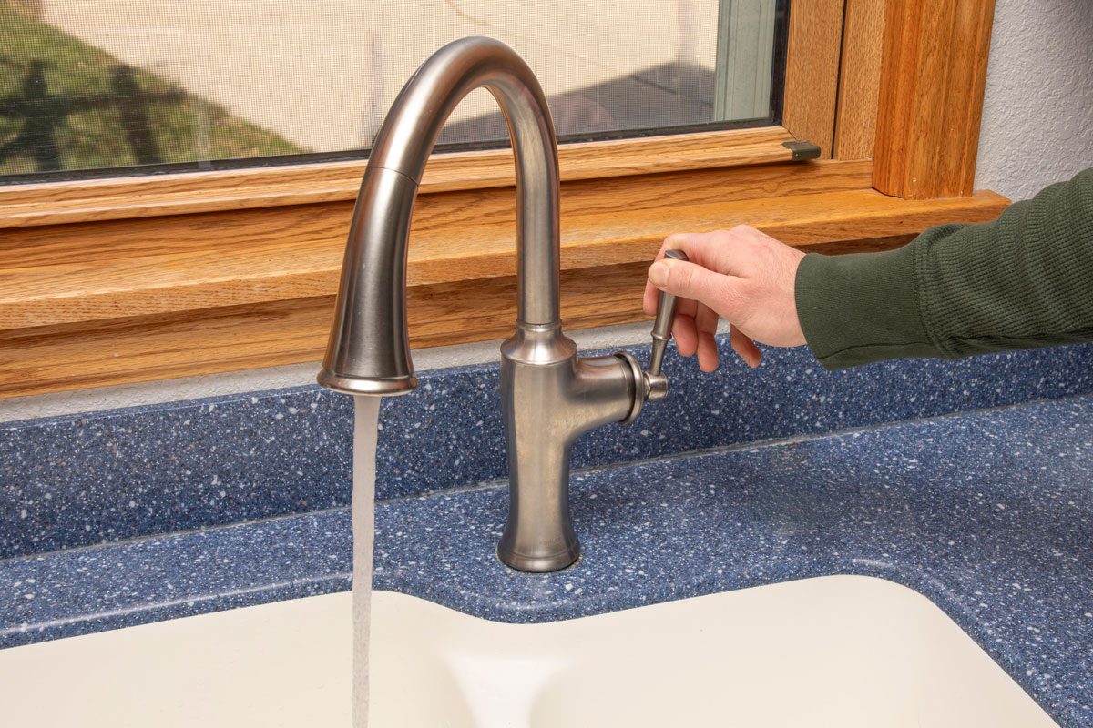 a hand turning the lever of a brushed metal kitchen faucet, and water is flowing out of the spout into a white sink; the faucet is mounted on a blue speckled countertop, and a wooden window frame with a screen is visible in the background