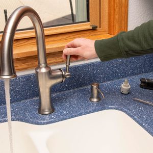 a brushed metal kitchen faucet with water running into a white sink; on the blue speckled countertop next to the faucet, there is a small metal cylinder with a chain, a white plastic cartridge, a black clamp like tool, a red and black screwdriver, and a metal wrench like tool; a hand is visible turning the faucet handle; a wooden window frame is in the background