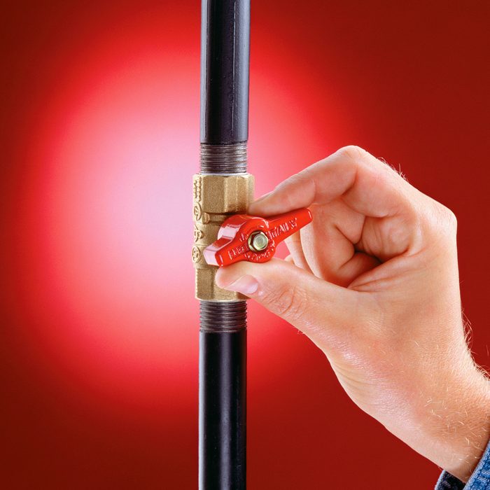 A hand turning a red valve handle on a brass pipe fitting attached to a vertical black pipe, with a red background.