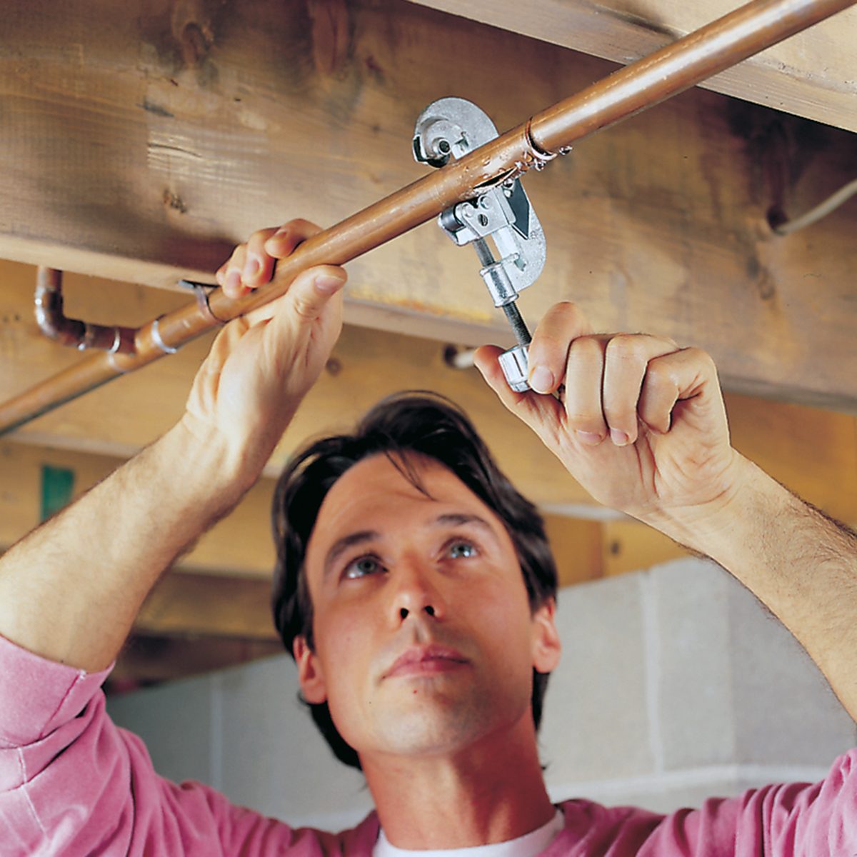 A man in a pink shirt uses a pipe cutter to cut a copper pipe attached to a wooden ceiling beam in a basement or utility space.